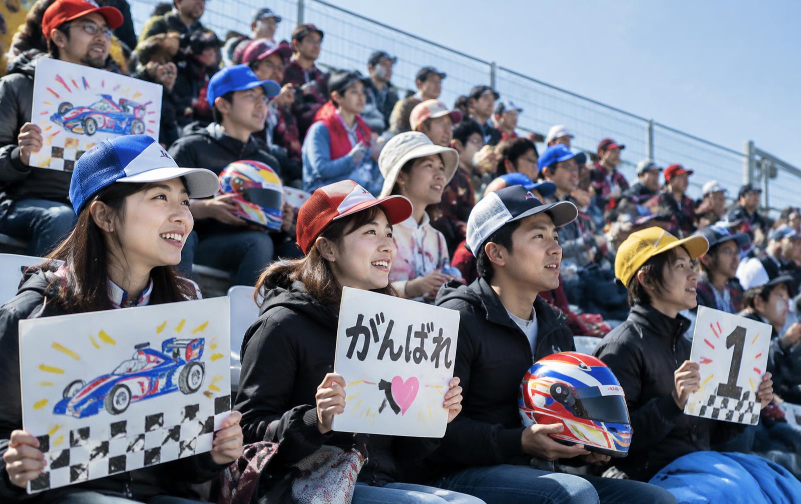 Motorsport fans in a circuit grandstand with handmade signs and replica helmets