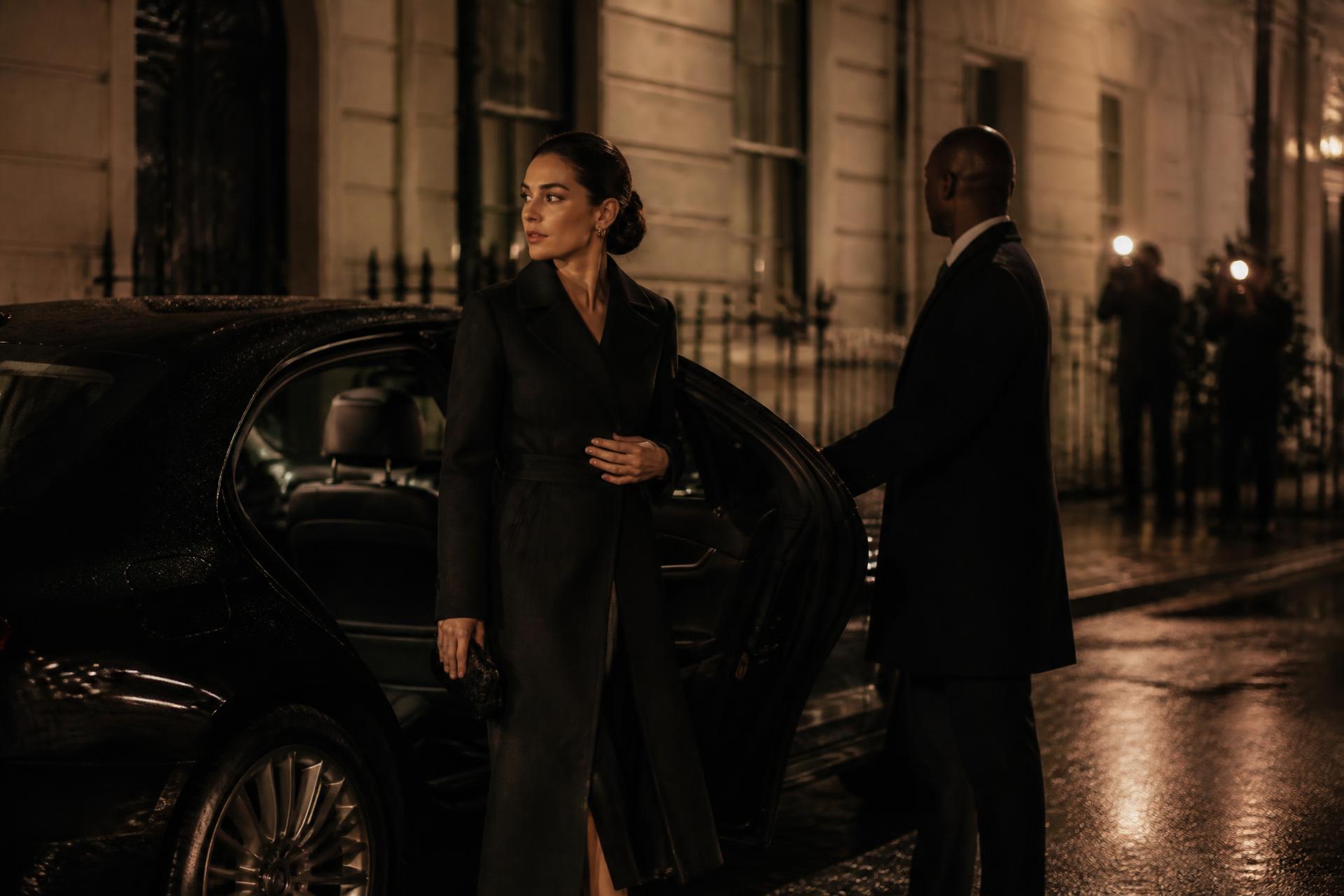 Guest in a long black coat stepping from a chauffeured car outside a lit stone London townhouse at night