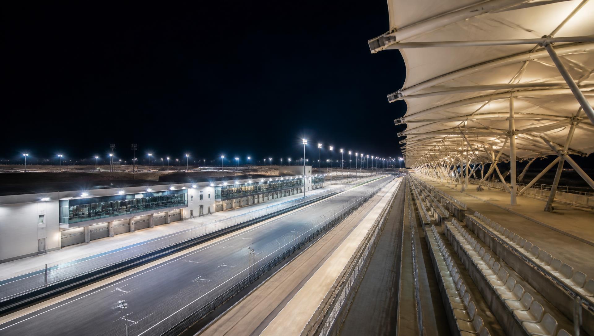 Floodlit main straight of a desert racing circuit with pit building and grandstand
