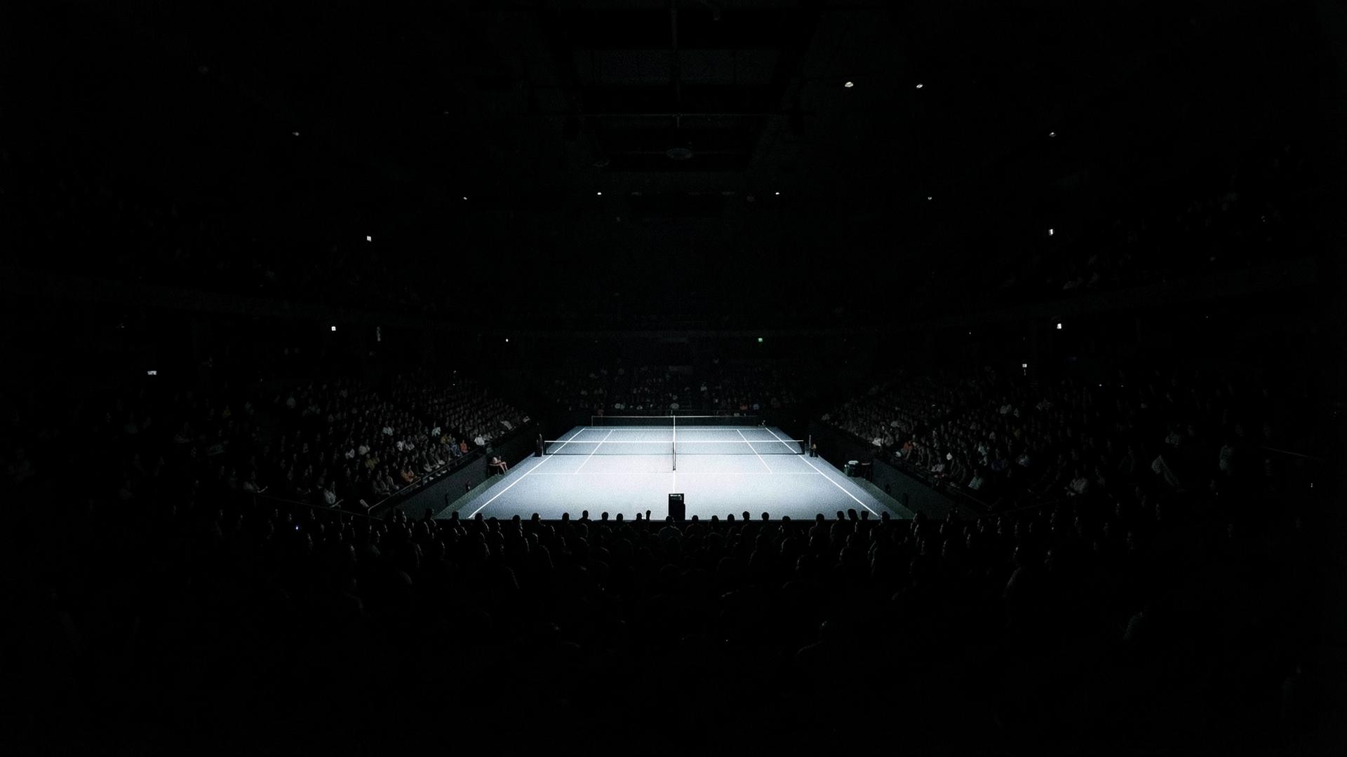 Single illuminated tennis court glowing inside an almost completely dark arena